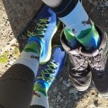Skarpetki długie z grafiką kozicy górskiej, na tle górskiej sceneri. Łąka z potokiem, białe szarotki i błękit nieba. Skarpetki ubrane do tregingowych butów, na kamienistym podłożu.