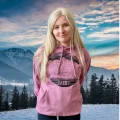 Morskie Oko Bluza Tatry Młoda, uśmiechnięta dziewczyna na tle zaśnieżonych szczytów. Ubrana w pudrową, ciepłą, bluzę z kapturem i dużą kieszenią z przodu. Grafika na ubraniu, przedstawia nowoczesne szczyty gór.