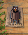 Haftowana broszka na drewnianym stole. Stojący na dwóch łapach, brązowy niedźwiedź brunatny. Jest przypięty do papierowej etykiety z napisem niedźwiedź i logo firmy.