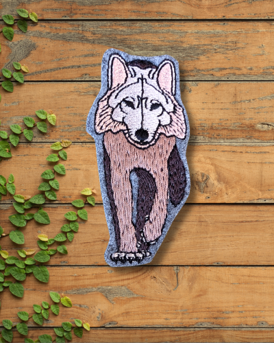 Haftowana broszka przedstawiająca samotnego, brązowego wilka. Wilk ma podniesioną przednią łapę, wygląda jak by dumnie kroczył. Zimne kolory, brąz, czerń i beż.
