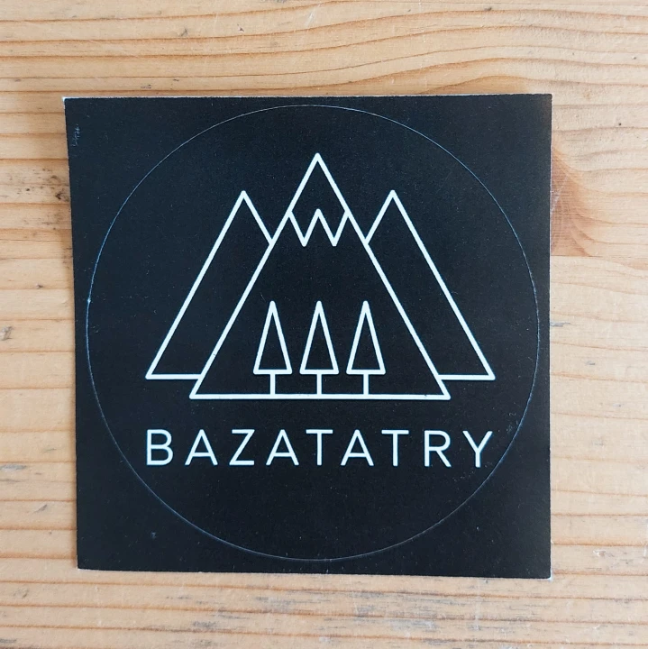 Czarna, kwadratowa naklejka z logo firmy. Biały kontur trzech szczytów, na dole trzy drzewka iglaste i napis bazatatry.