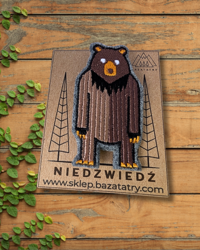 Haftowana broszka na drewnianym stole. Stojący na dwóch łapach, brązowy niedźwiedź brunatny. Jest przypięty do papierowej etykiety z napisem niedźwiedź i logo firmy.