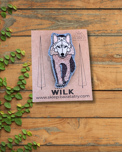 Haftowana broszka przedstawiająca samotnego, brązowego wilka. Wilk ma podniesioną przednią łapę, wygląda jak by dumnie kroczył. Zimne kolory, brąz, czerń i beż. Papierowa etykieta z logo firmy.