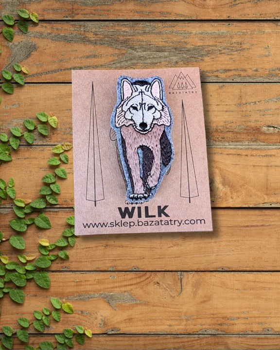 Haftowana broszka przedstawiająca samotnego, brązowego wilka. Wilk ma podniesioną przednią łapę, wygląda jak by dumnie kroczył. Zimne kolory, brąz, czerń i beż. Papierowa etykieta z logo firmy.